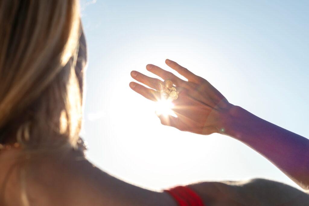 A person with blonde hair holds their hand up to the bright blue sky. The sunlight shines through their fingers and makes a butterfly ring sparkle, creating a peaceful mood.
