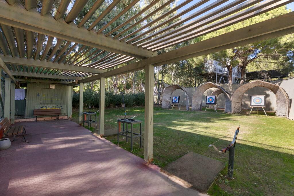 Covered archery range at the all-inclusive Megasaray Club Belek resort with three targets on a grassy area. A wooden pergola provides shade and creates a relaxed atmosphere on sunny days. The bows are stored on racks.