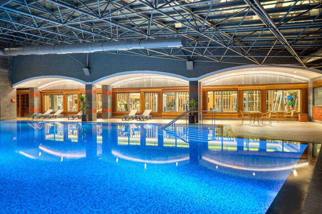 Modern indoor pool at the GSTC-certified Megasaray Club Belek resort with sparkling blue water, surrounded by wooden deck chairs and potted plants. The ceiling features exposed metal beams.