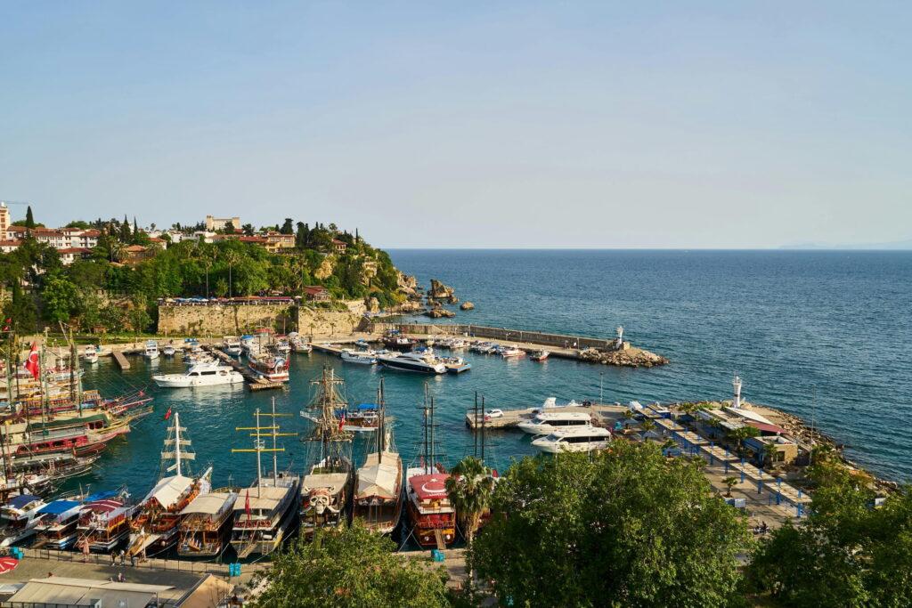 Beautiful view of the coastline of a harbor in Antalya, Turkey, with many boats and yachts moored there. The area is surrounded by lush greenery and cliffs, with the calm, vast blue sea stretching out behind it.
