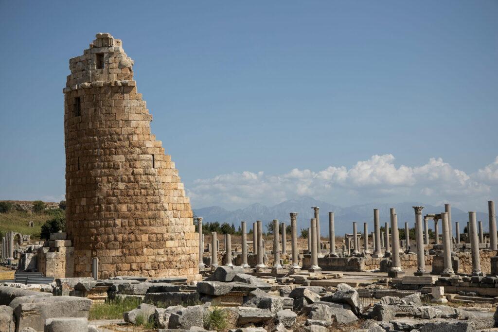 Ancient ruins of a round tower and columns under a bright blue sky near Antalya, Turkey. The stone structures convey a sense of history and archaeological significance.