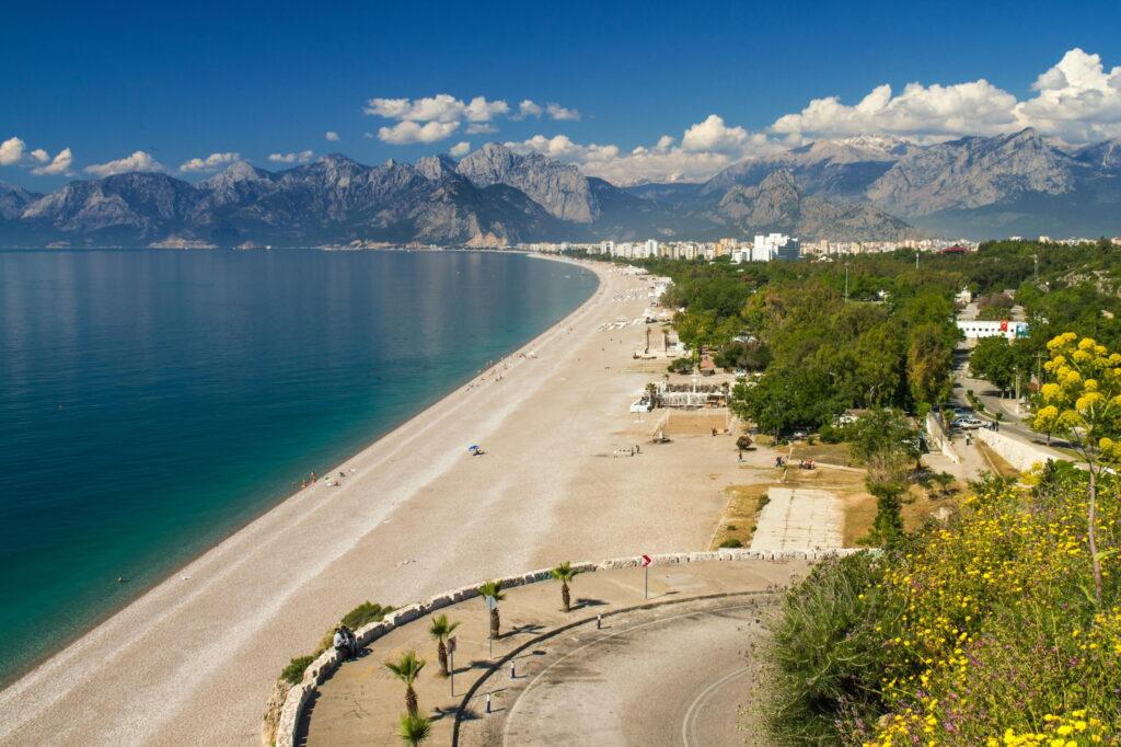 A stunning view of the coast near Antalya in Turkey shows a long, curved beach with golden sand bordering turquoise waters. Lush greenery and mountains in the distance add to the beauty of this tranquil setting.