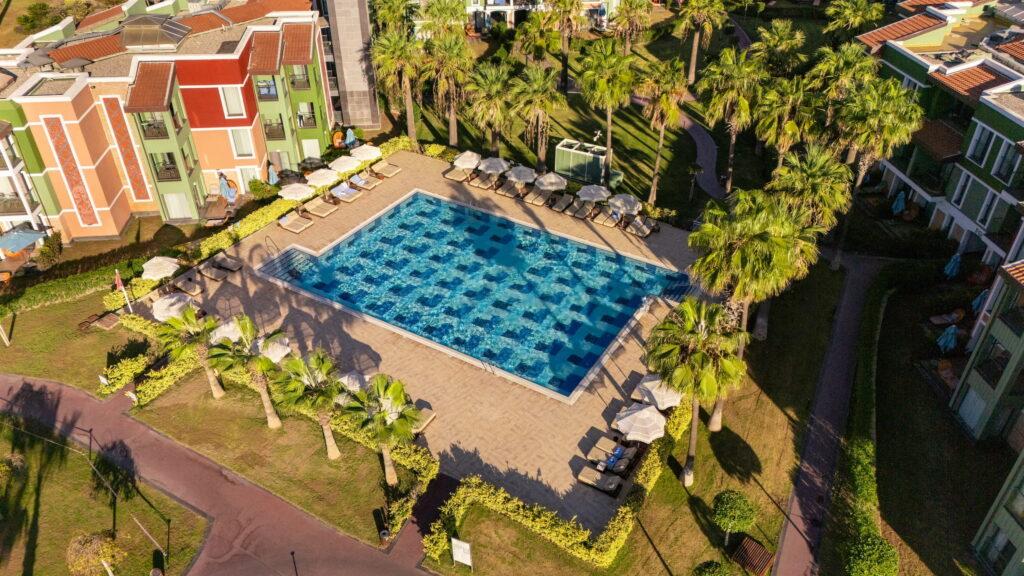 Aerial view of a rectangular pool at the all-inclusive Megasaray Club Belek resort, surrounded by sun loungers and parasols, with palm trees and colorful buildings in the background.