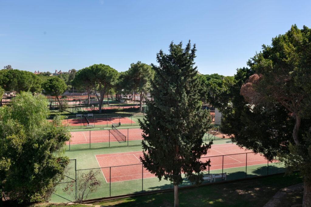 The tennis courts at the GSTC-certified all-inclusive resort Megasaray Club Belek are surrounded by tall green trees and set beneath a bright blue sky, creating a peaceful and inviting atmosphere for outdoor sports.