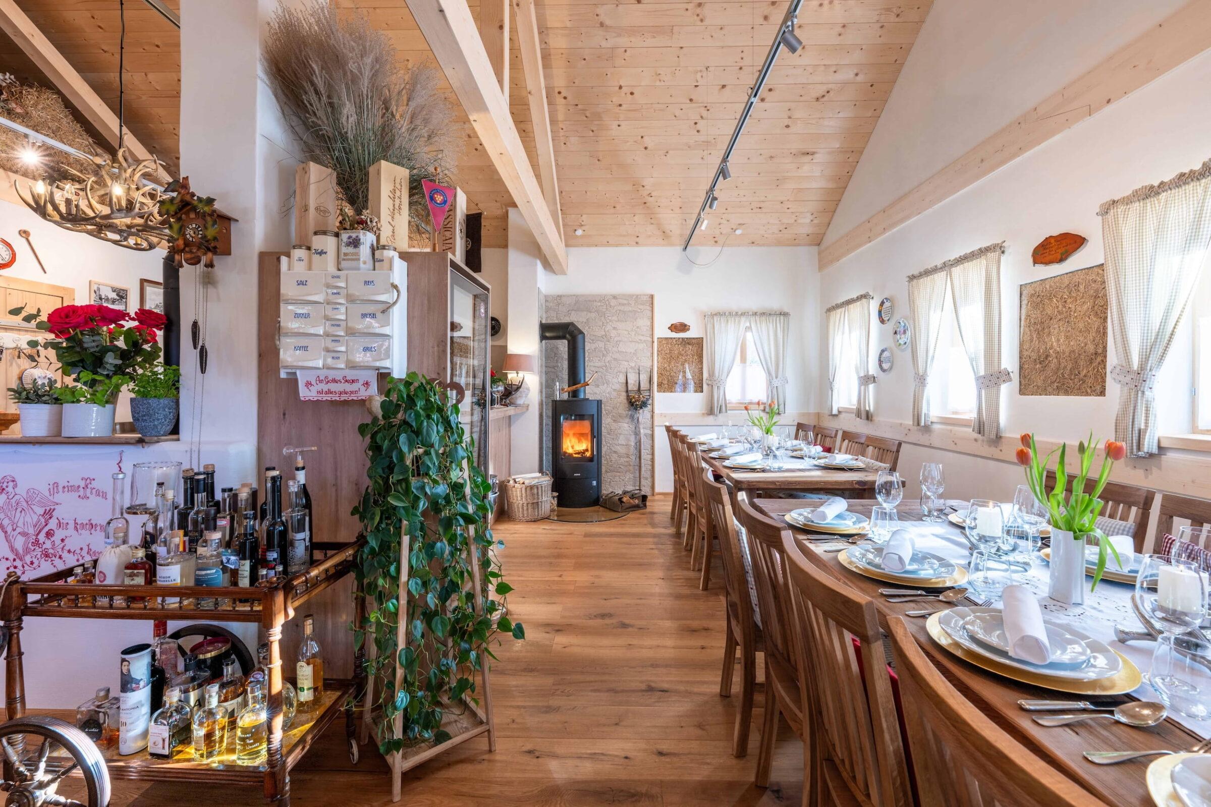 Spacious rustic dining room in the “Dorfschenke” at Ebner 1822 in Carinthia with a long wooden table set for guests. Warm, cozy atmosphere with a fireplace, plants, and decorative shelves.