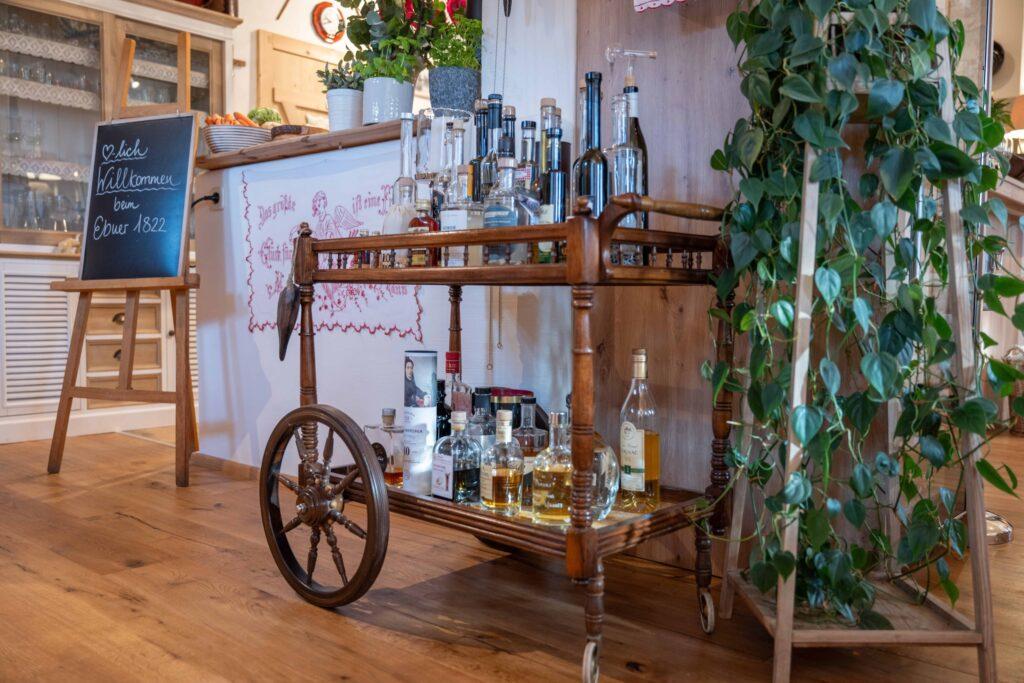A wooden cart with various bottles stands in the cozy Dorfschenke restaurant at Ebner 1822 next to a lush potted plant and an inviting table on an easel. Warm, inviting atmosphere.