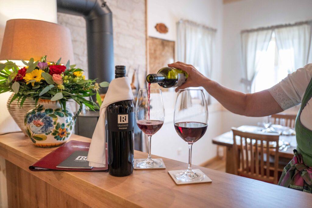 Cozy restaurant Dorfschenke von Ebner 1822 in Carinthia with a person pouring red wine into two glasses on a wooden counter. Nearby are a colorful vase of flowers and a bottle of wine.