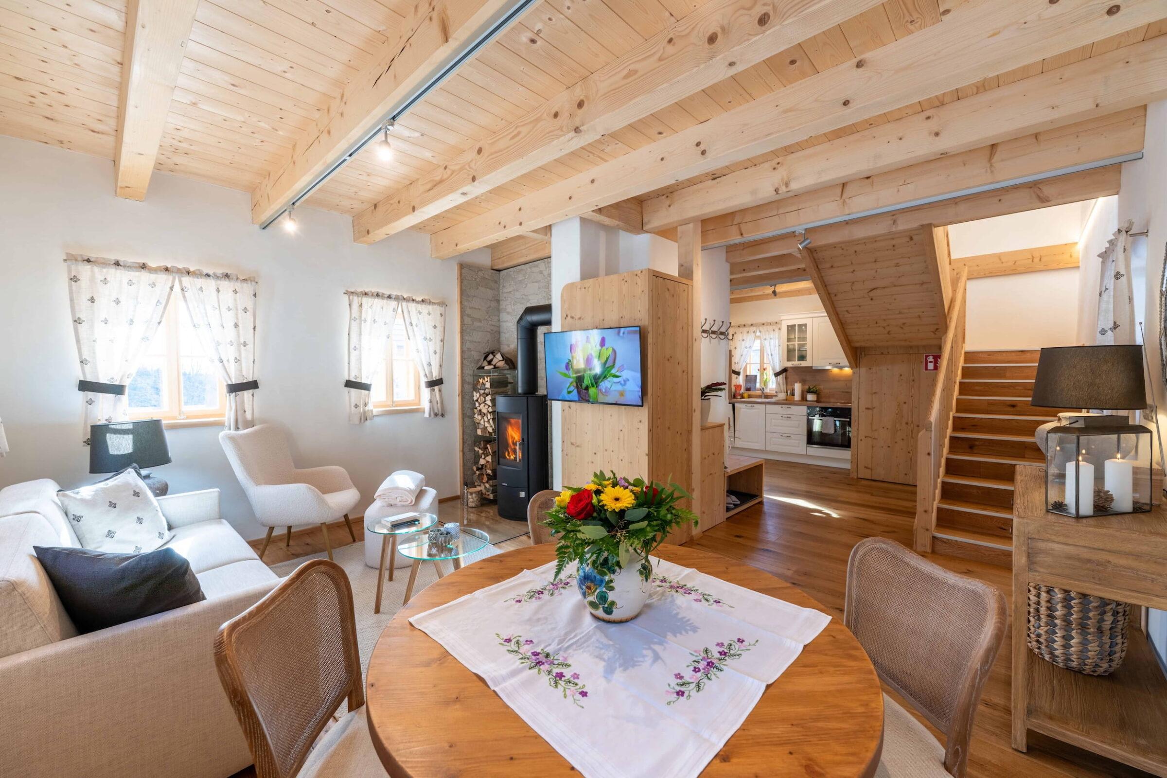 Cozy wooden interior of a living room in the Ebner 1822 vacation homes in Carinthia, Austria, with a sofa, armchairs, TV, and fireplace. In front of it stands a dining table with flowers, radiating warmth.