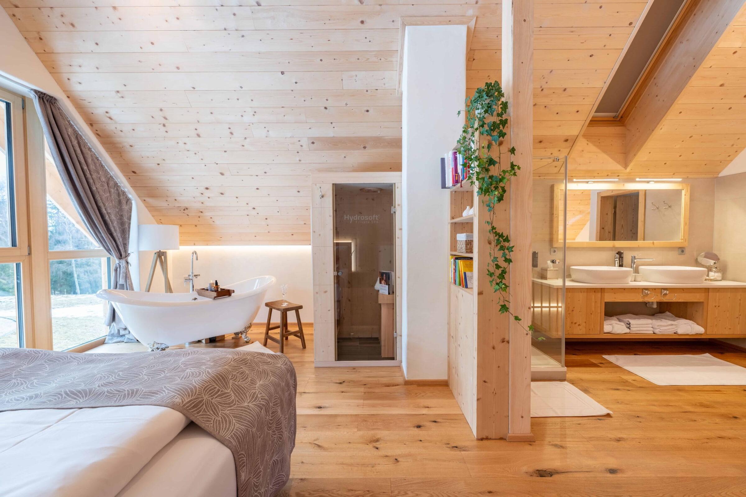 Spacious loft bedroom with wood paneling, freestanding bathtub near large windows, cozy double bed, sauna, and double sink in the Ebner 1822 vacation homes in Carinthia, Austria. Relaxing atmosphere.