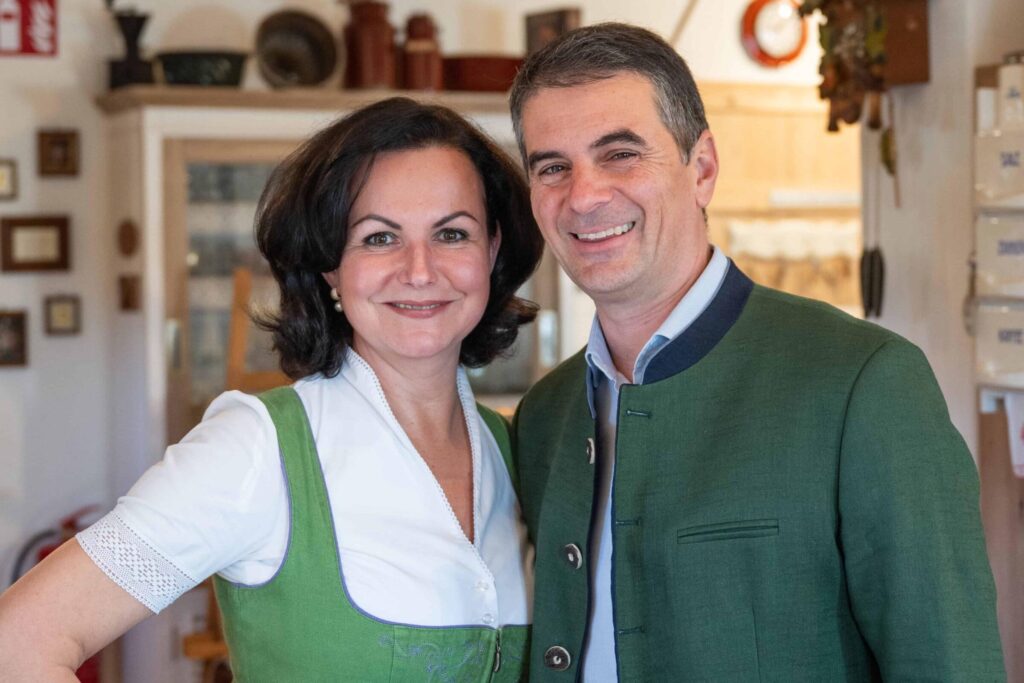 Ein Mann und eine Frau, die Eigentümer der Ferienwohnungen Ebner 1822 in Österreich, lächeln in Innenräumen und tragen traditionelle österreichische Trachten. Die Frau trägt ein grünes Dirndl mit Schürze, der Mann eine grüne Jacke. Gemütliche und einladende Atmosphäre.