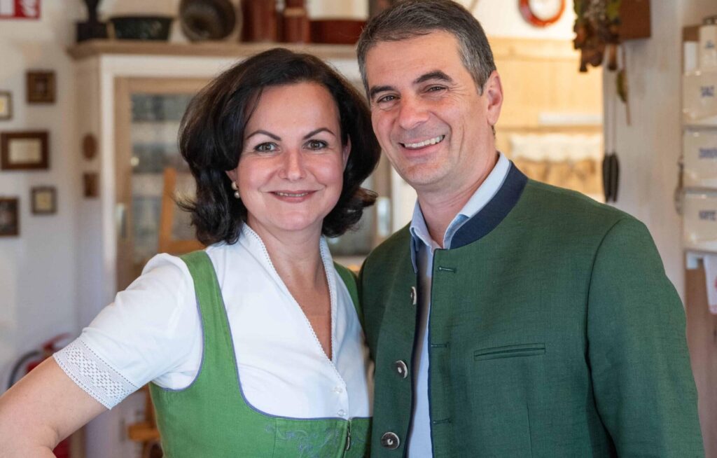 A man and a woman, the owners of Ebner 1822 vacation rentals in Austria, smile indoors wearing traditional Austrian costumes. The woman wears a green dirndl with an apron, and the man wears a green jacket. Cozy and inviting atmosphere.