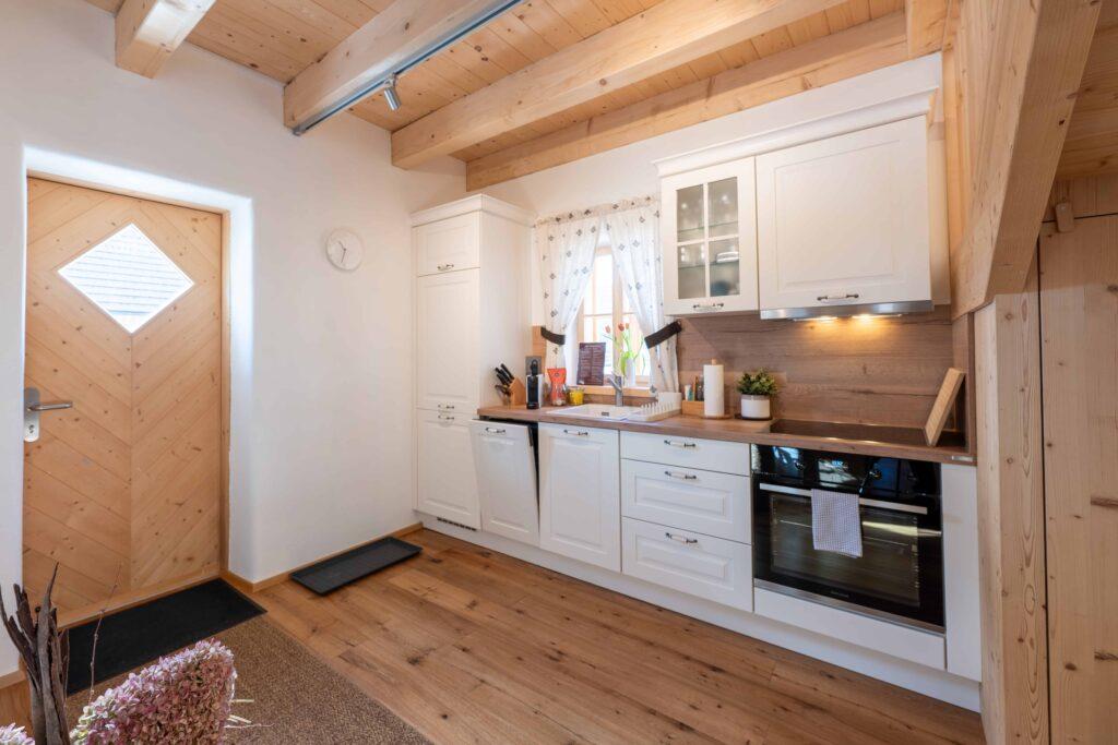 A cozy kitchen in one of the Ebner 1822 vacation homes in Austria with wooden beams and wooden floors. White cabinets are accented by a wooden countertop. A curtain hangs over a small window and radiates warmth.