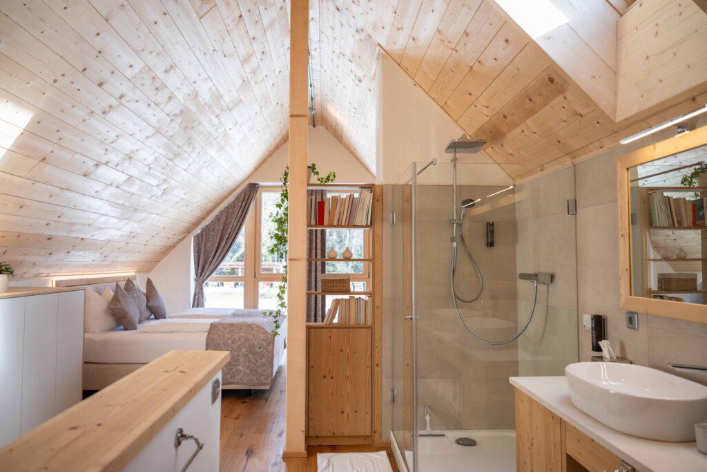 Cozy bedroom in the attic of one of the Ebner 1822 vacation homes in Carinthia with wooden ceiling and wood paneling. On the left is a bed with gray pillows, on the right is a modern bathroom with a glass shower. Natural light streams in through a window with beige curtains, creating a warm, inviting atmosphere.