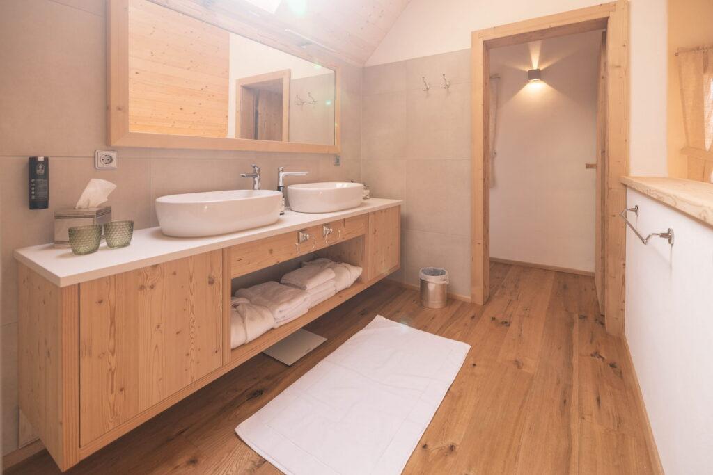 The tranquil, modern bathroom in the Ebner 1822 vacation apartments in Carinthia features wood accents and has a double vanity with two white sinks, a large mirror, a towel rack, and soft lighting.