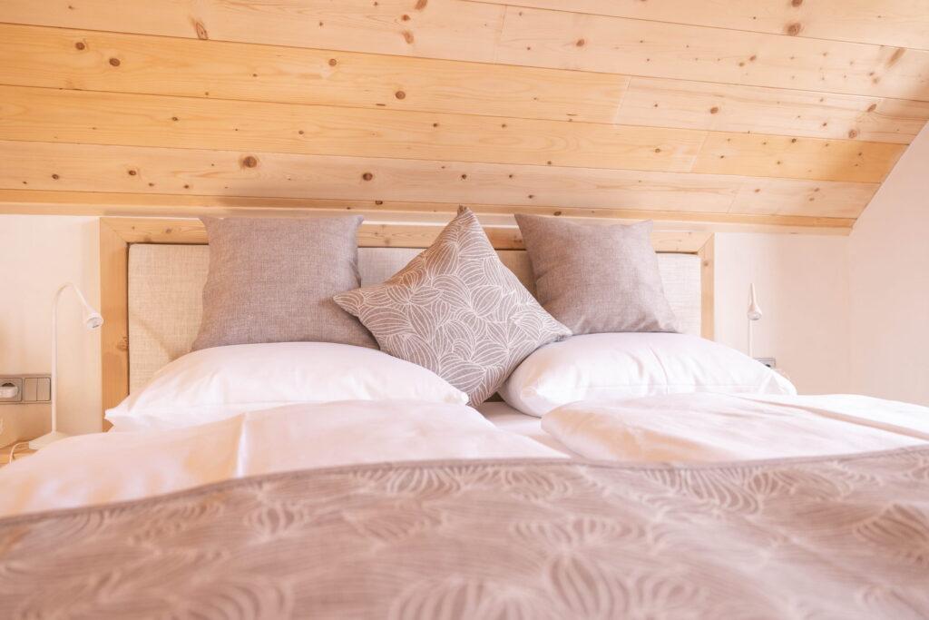 Cozy bedroom in one of the three vacation homes at Ebner 1822 with a neatly made bed, three decorative gray pillows, and a patterned beige bedspread. The sloping wooden ceiling adds to the cozy atmosphere.