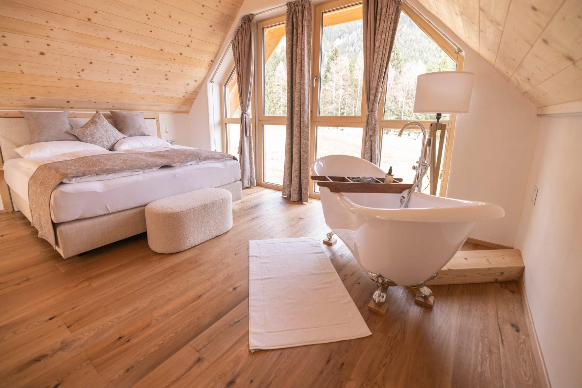 Cozy attic room in an Ebner 1822 vacation home with a double bed, white bedding, and a freestanding bathtub near large windows. Warm wood tones create a calm, inviting atmosphere.