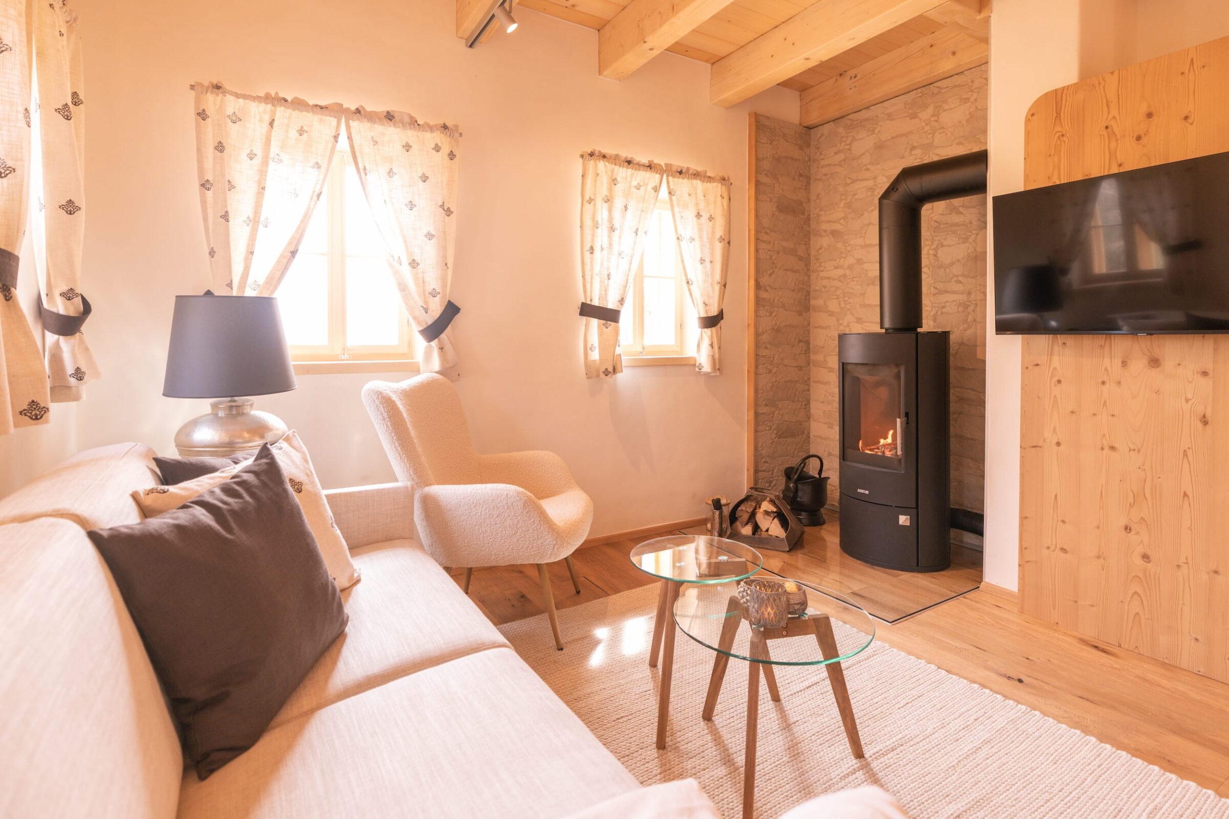 Cozy living room at Ebner 1822 in Austria with a beige sofa, an armchair, a wood-burning stove, and a glass coffee table. The warm lighting from two windows creates a peaceful atmosphere.