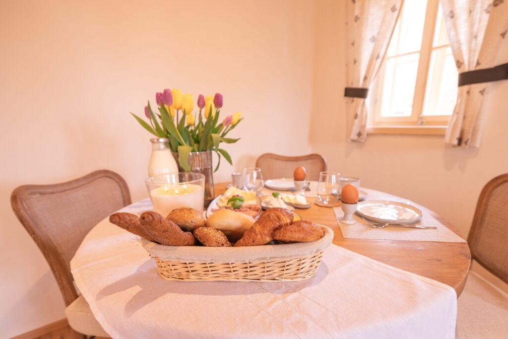 A cozy table set for an organic breakfast at the Ebner 1822 vacation apartments in Carinthia, with a basket full of different types of bread and baked goods and bright tulips in a vase. Soft daylight streams through the window, creating a warm, inviting atmosphere.