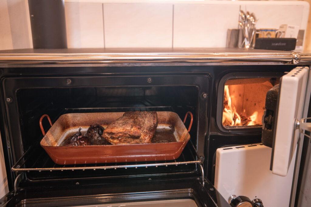 Organic roast beef in a rectangular dish in an open oven, whose flames are visible in a side chamber. The atmosphere is warm and rustic in the kitchen of the Ebner 1822 Dorfschenke in Carinthia.