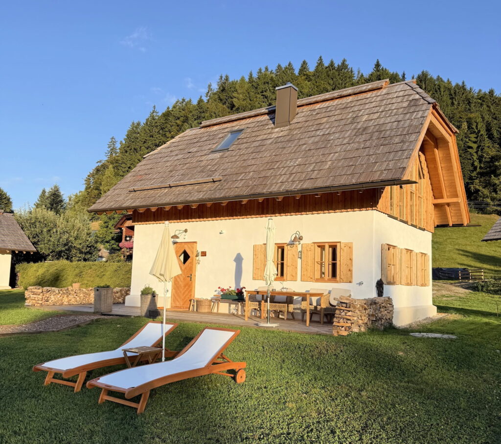 A quaint, rustic vacation home built by Ebner in 1822 in Austria with wooden shutters and a sloping roof is surrounded by greenery. Two sun loungers stand on the lawn, creating a peaceful, relaxed atmosphere.