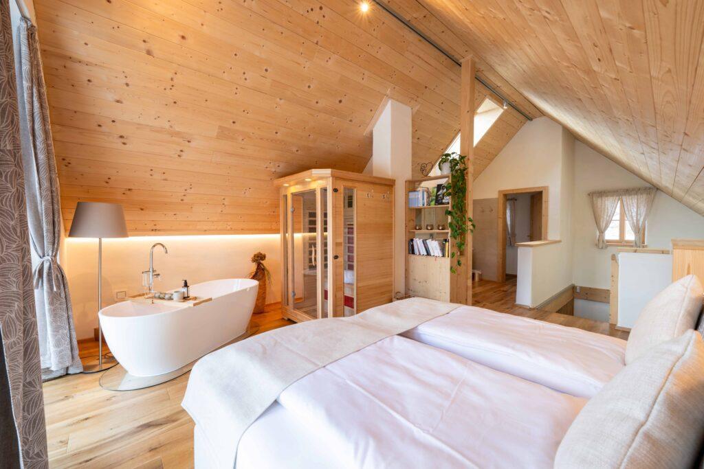 Cozy attic bedroom with wooden walls and floors in the Ebner 1822 vacation homes in Carinthia, Austria. Featuring a white freestanding bathtub, a soft bed, a large lamp, and a potted plant that exude a calm, spa-like atmosphere.