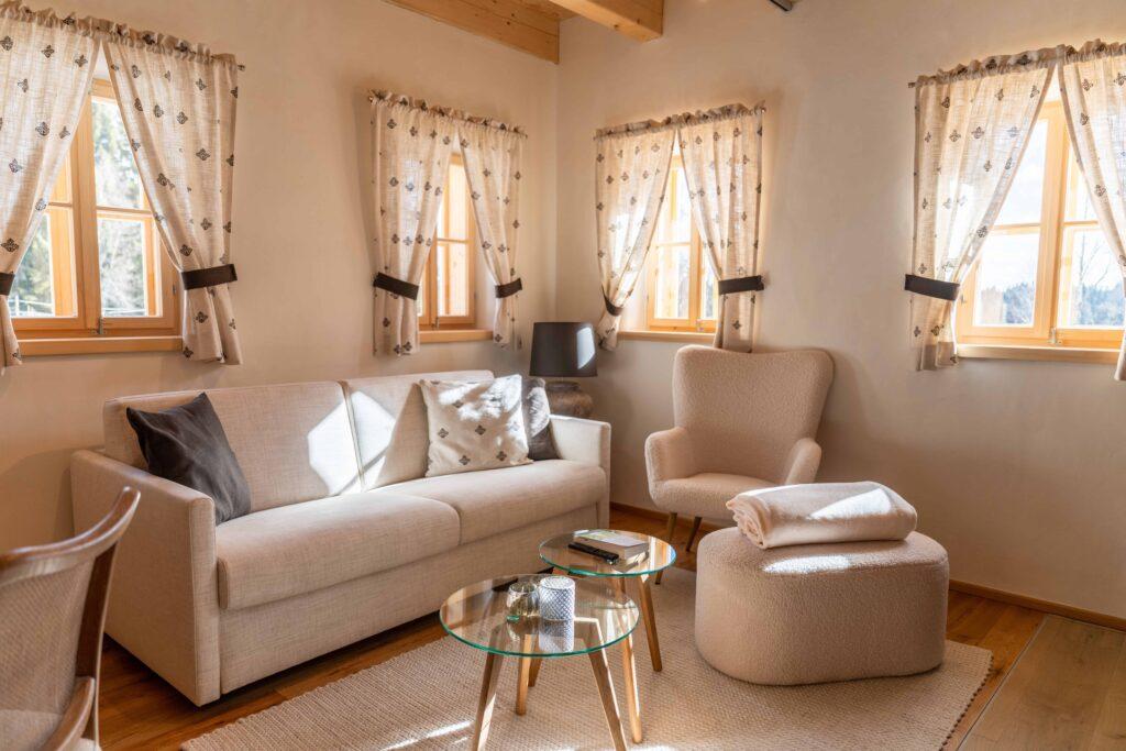 Bright and cozy living room in Ebner 1822's Austrian vacation homes, with a beige sofa, armchair, and round glass coffee table. Sunlight streams in through four windows with patterned curtains.