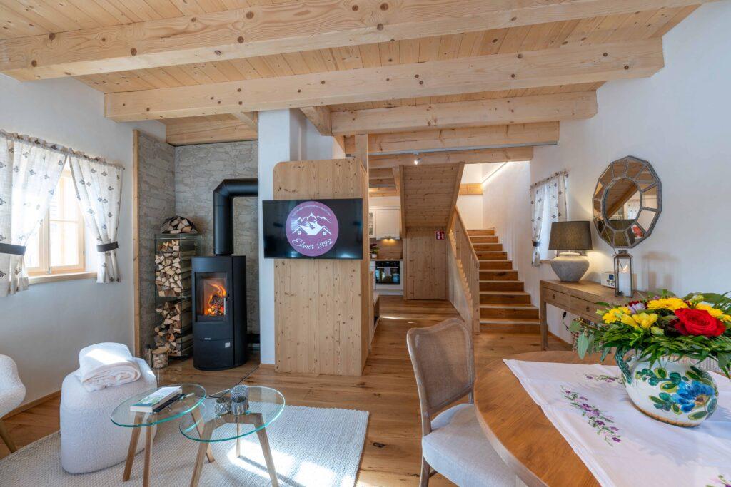 Cozy living room with wooden beams and wooden floors in the Ebner 1822 vacation home in Carinthia, Austria. With a fireplace and a TV. A staircase leads upstairs. Bright flowers adorn the dining table. Inviting and warm.