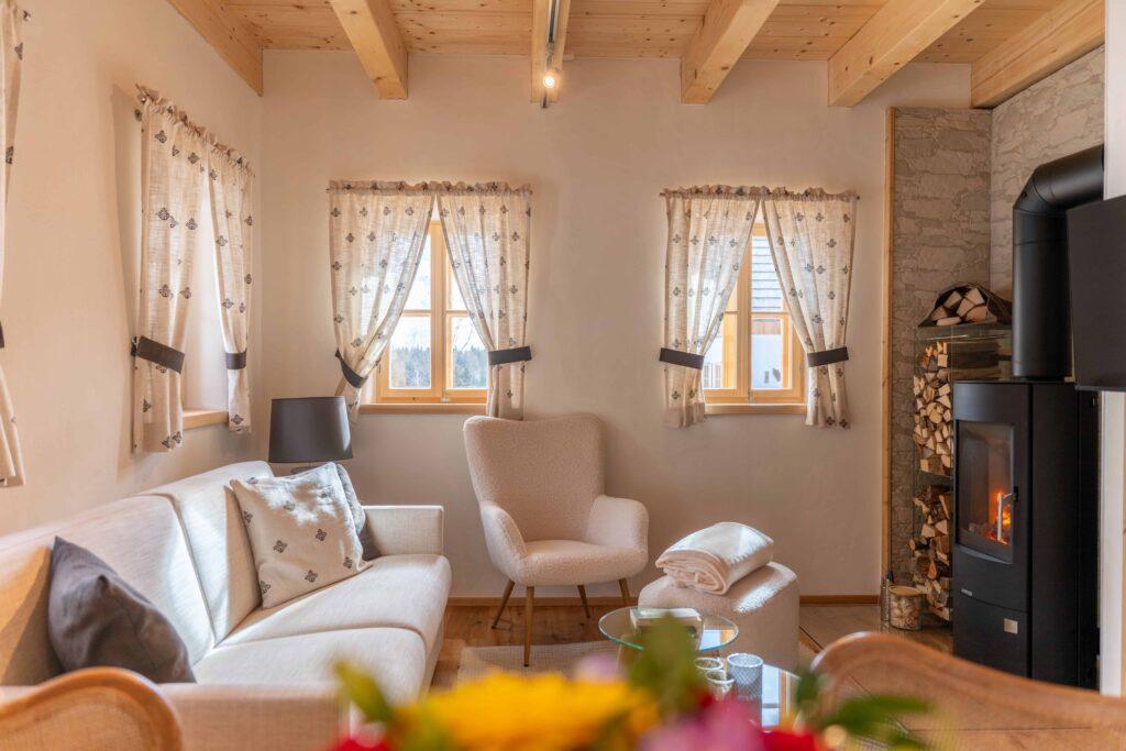 Cozy living room in one of Ebner 1822's vacation homes in Carinthia, Austria, with a beige sofa, armchair, and wood-burning stove. Two windows with patterned curtains let in natural light. Warm, inviting atmosphere.