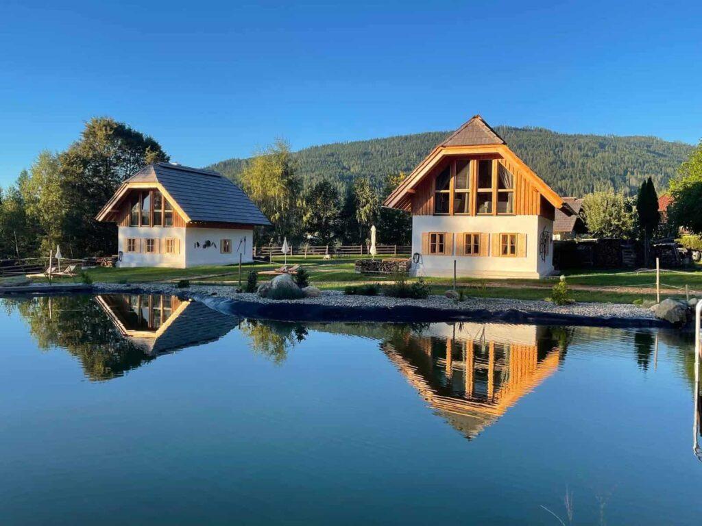 Two charming wooden holiday homes by Ebner 1822 in Austria with sloping roofs and large windows are reflected in a tranquil pond, set against a backdrop of lush trees and a clear blue sky.