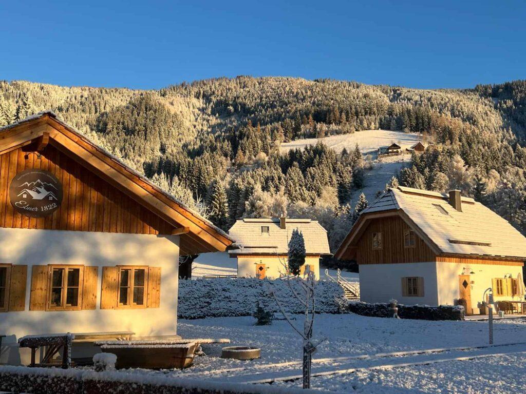 Snow-covered wooden chalets from Ebner 1822 in Carinthia, Austria, located in a sunny, quiet valley against a backdrop of pine-covered mountains, exude a peaceful, wintery charm.