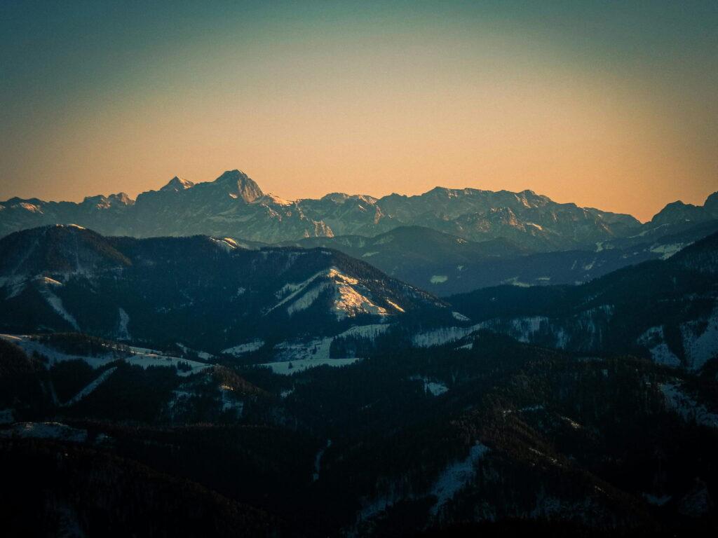 Sunset over a mountain range in Carinthia, Austria, with snow-capped peaks under a warm, orange sky. Majestic and tranquil atmosphere that conveys serenity.