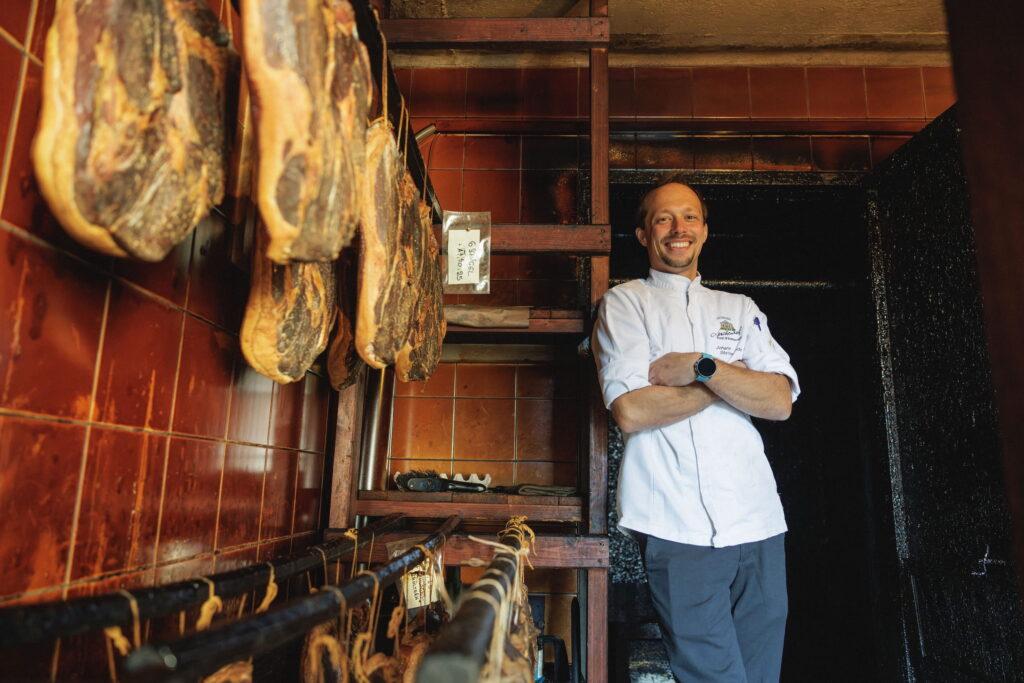 Chef Johann from the sustainable Hotel Schloss Lerchenhof stands smiling in his white uniform in a room where sausages hang from hooks. The decor is rustic, with warm brown tiled walls.