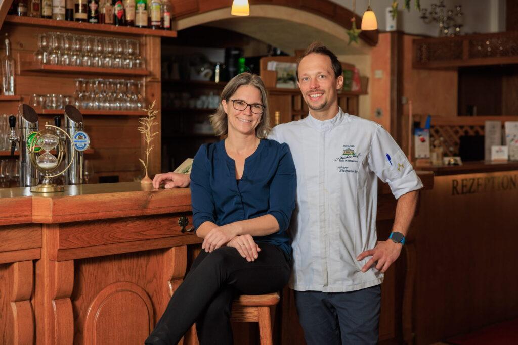 The hostess of the Hotel Schloss Lerchenhof, Karin Steinwender, wearing a blue blouse, sits on a bar stool next to her partner and owner Johann, who is wearing a chef's uniform. They are in the cozy, dimly lit bar surrounded by glasses and bottles. Both are smiling.