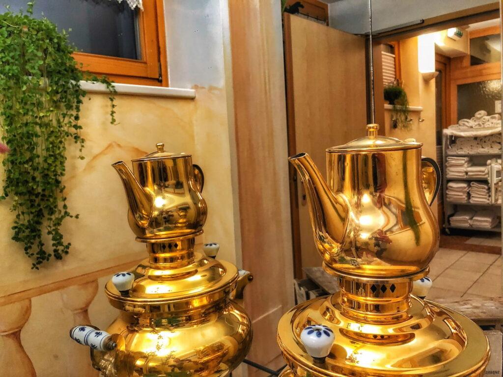 A golden samovar with ornate handles is reflected in the mirror in the wellness area of the sustainable Hotel Schloss Lerchenhof in Carinthia. A lush green plant hangs nearby, and towels are neatly stacked in the softly lit room.