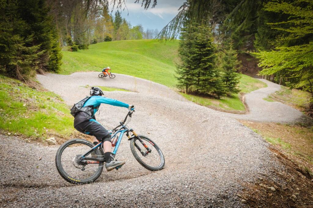 Two mountain bikers ride along a winding gravel path through a lush forest area in Carinthia, Austria. The rider in front leans into a curve, embodying excitement and a spirit of adventure.