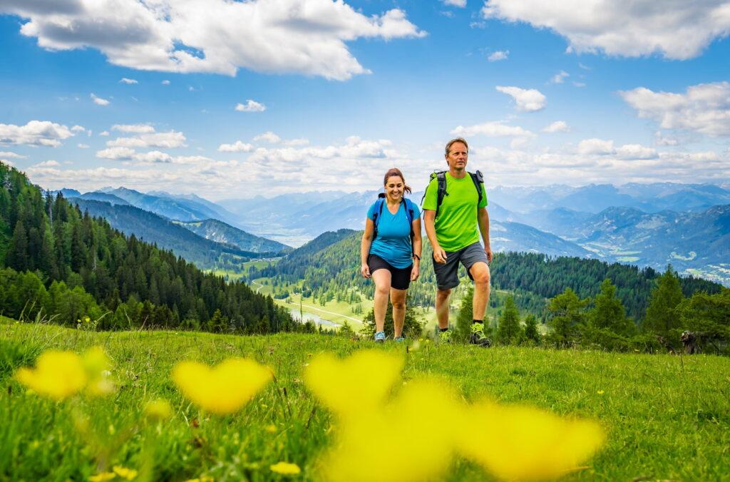 A man and a woman hike up a grassy mountainside in Carinthia, Austria, under a blue sky with clouds. The picturesque view shows lush green forests and distant mountains, conveying a sense of adventure and tranquility. Yellow flowers are in the foreground.