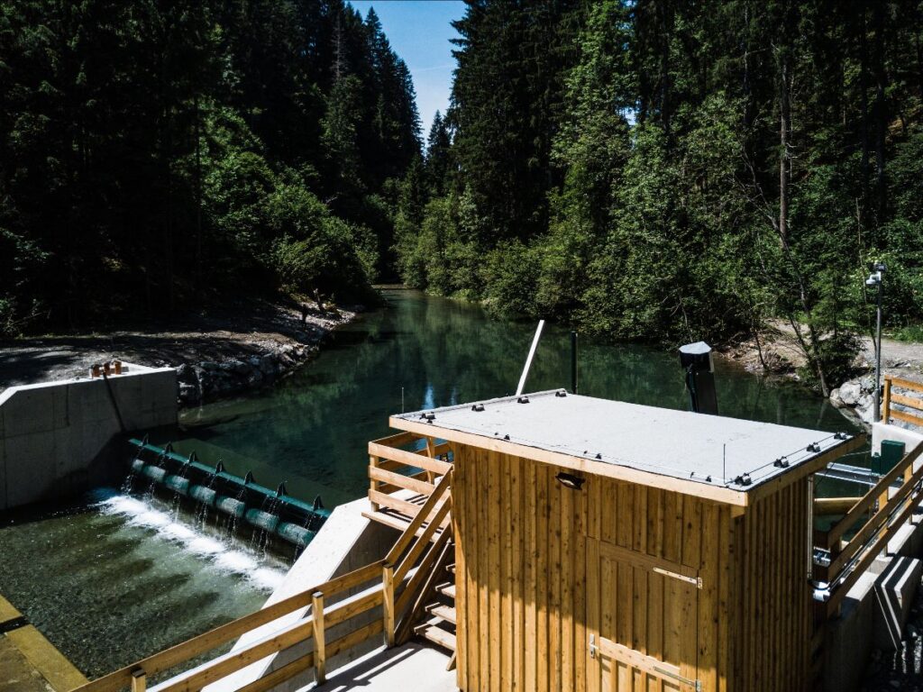A small hydroelectric dam in the middle of a forest, with a wooden control building in the foreground, supplies the sustainable Hotel Schloss Lerchenhof in Carinthia, Austria. The calm river and dense trees create a peaceful atmosphere.