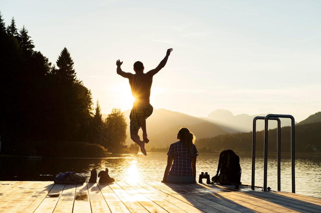One person happily jumps from a jetty into a sun-drenched lake in Carinthia, Austria, while another person watches the scene. The sunlight glitters on the water, surrounded by trees and distant hills.