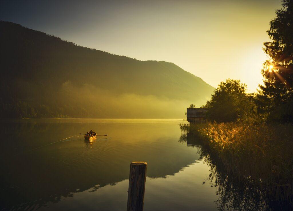 A tranquil lake scene in Austria at sunrise, with a person rowing a boat on the calm water. Fog-covered hills and a bright sun create a peaceful, golden atmosphere.