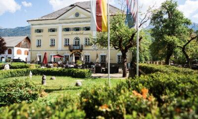 Das historische gelbe Gebäude des nachhaltigen Hotels Schloss Lerchenhof mit symmetrischen Fenstern und einem Balkon, umgeben von gepflegten Gärten und Fahnen. Ein strahlender, sonniger Tag.