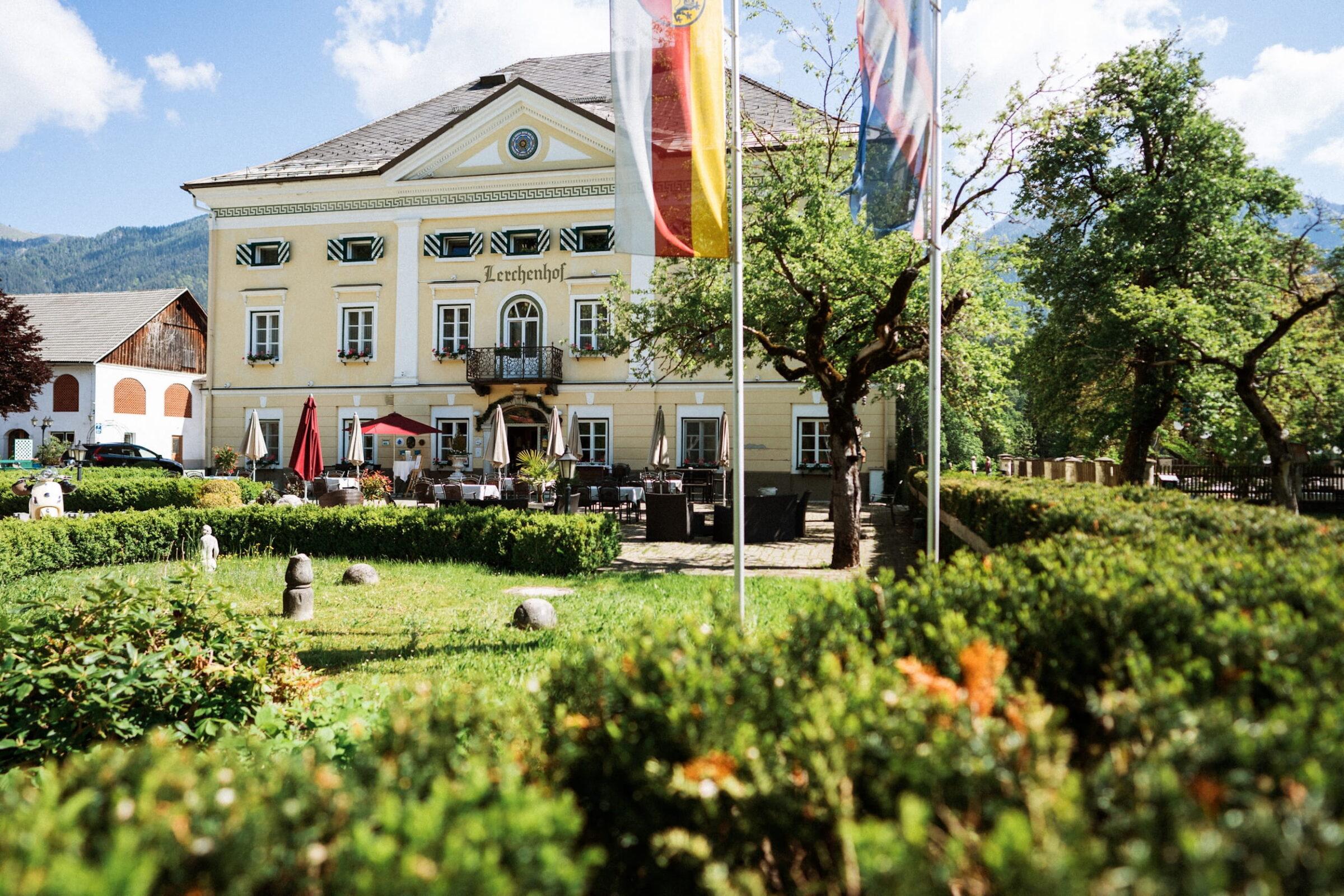 The historic yellow building of the sustainable Hotel Schloss Lerchenhof with symmetrical windows and a balcony, surrounded by well-tended gardens and flags. A bright, sunny day.