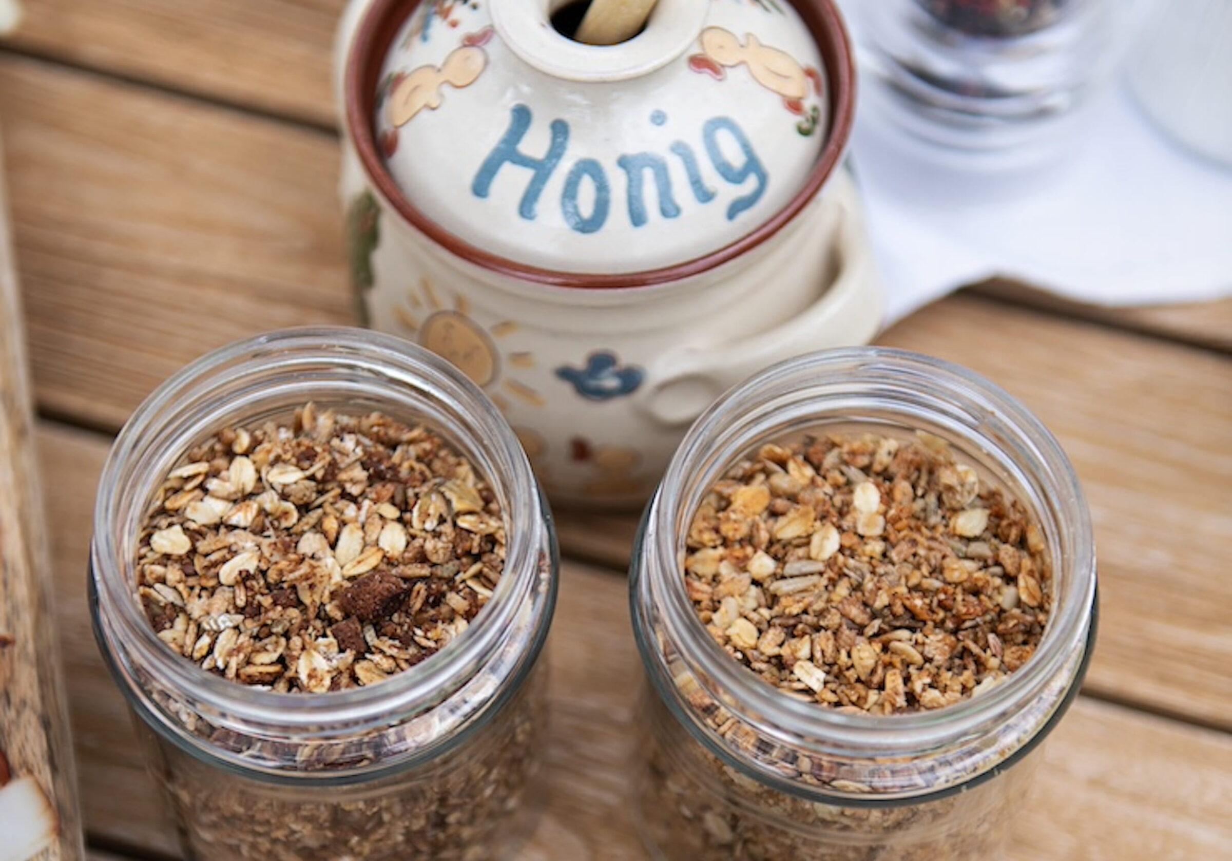 Two jars filled with organic muesli stand on a wooden table next to a ceramic honey jar labeled “Honey,” creating a rustic, cozy breakfast scene from the sustainable Hotel Schloss Lerchenhof.