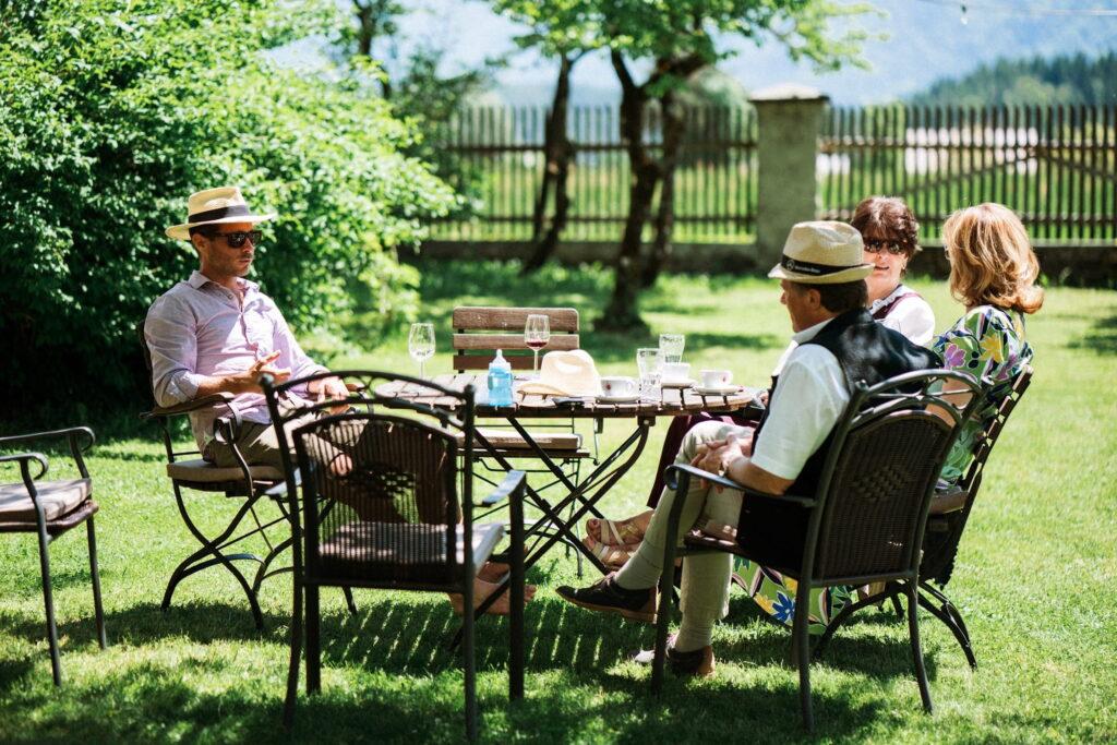 Four people are sitting around a garden table in the castle garden of the Hotel Schloss Lerchenhof on a sunny day, wearing summer hats and casual clothes. They are surrounded by lush greenery and blue skies.