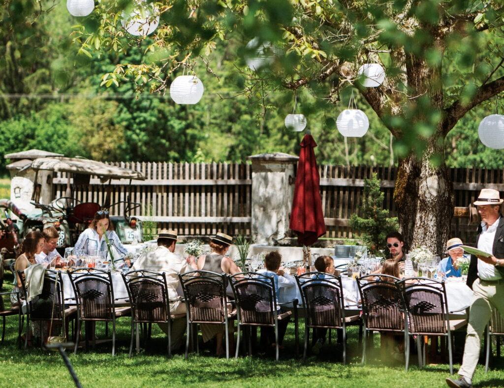 Garden wedding at the sustainable Hotel Schloss Lerchenhof with people sitting at a long table under a tree. Lanterns hang above them, creating a festive, relaxed atmosphere. Sunlight filters through the leaves.
