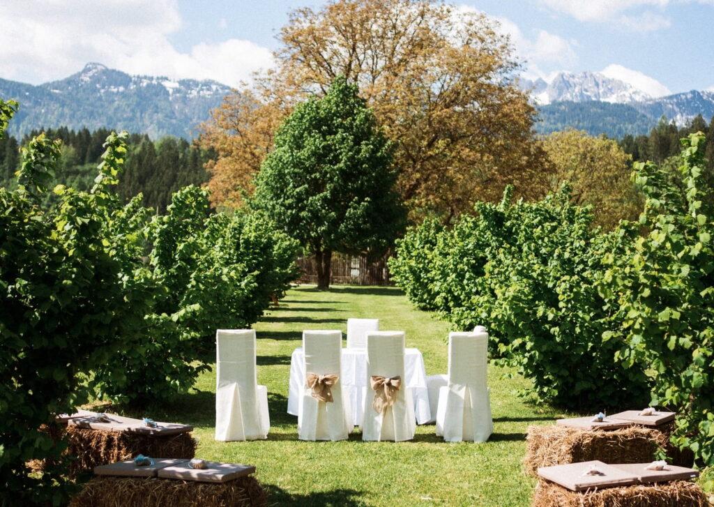 An outdoor wedding at Lerchenhof Castle with white chairs decorated with ribbons, hay bales with cushions, between rows of green trees and picturesque mountains in the background.