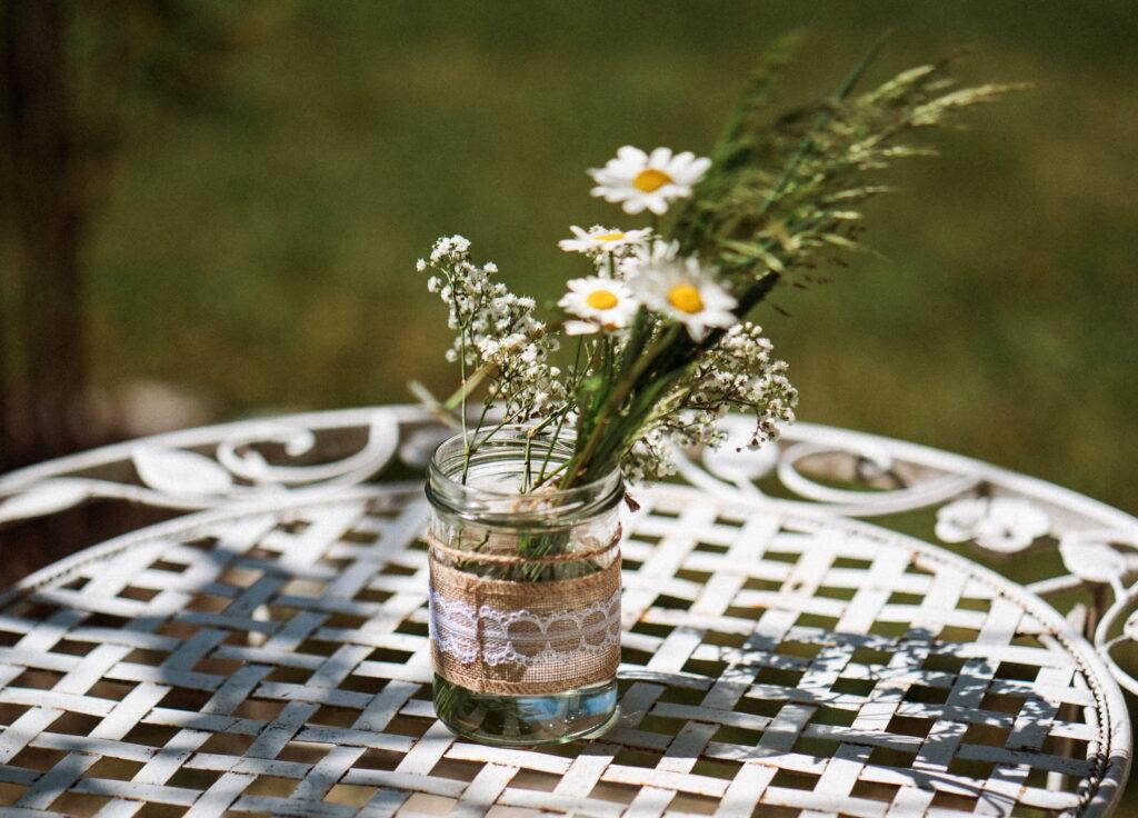 A glass vase filled with daisies and green plants, decorated with lace and burlap, stands on a white lattice table in the garden of the sustainable boutique hotel Schloss Lerchenhof. The sun-drenched scene conveys a sense of tranquility and rurality.