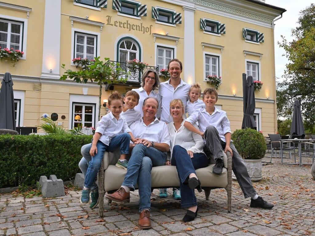The Steinwender family, owners of the sustainable Hotel Schloss Lerchenhof in Austria, pose in front of a yellow building with green shutters, all wearing white shirts and jeans. Smiling and relaxed, they convey warmth and togetherness.