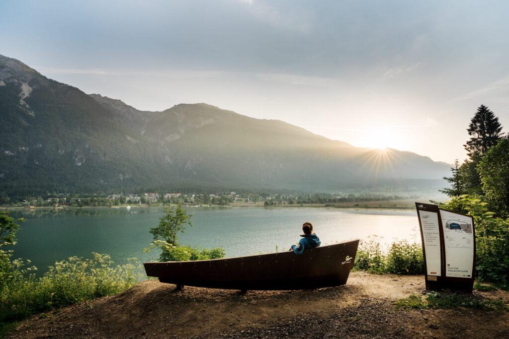A person sits on a boat-shaped bench on the Slow Trail near the Hotel Schloss Lerchenhof, gazing out at a tranquil lake and the mountains at sunrise. Soft light underscores the peaceful, contemplative scene.