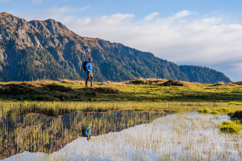 A solitary hiker in a blue jacket stands on a grassy ridge in Carinthia, Austria, with a mountainous landscape stretching out behind him. A reflective pond in the foreground mirrors the scene.