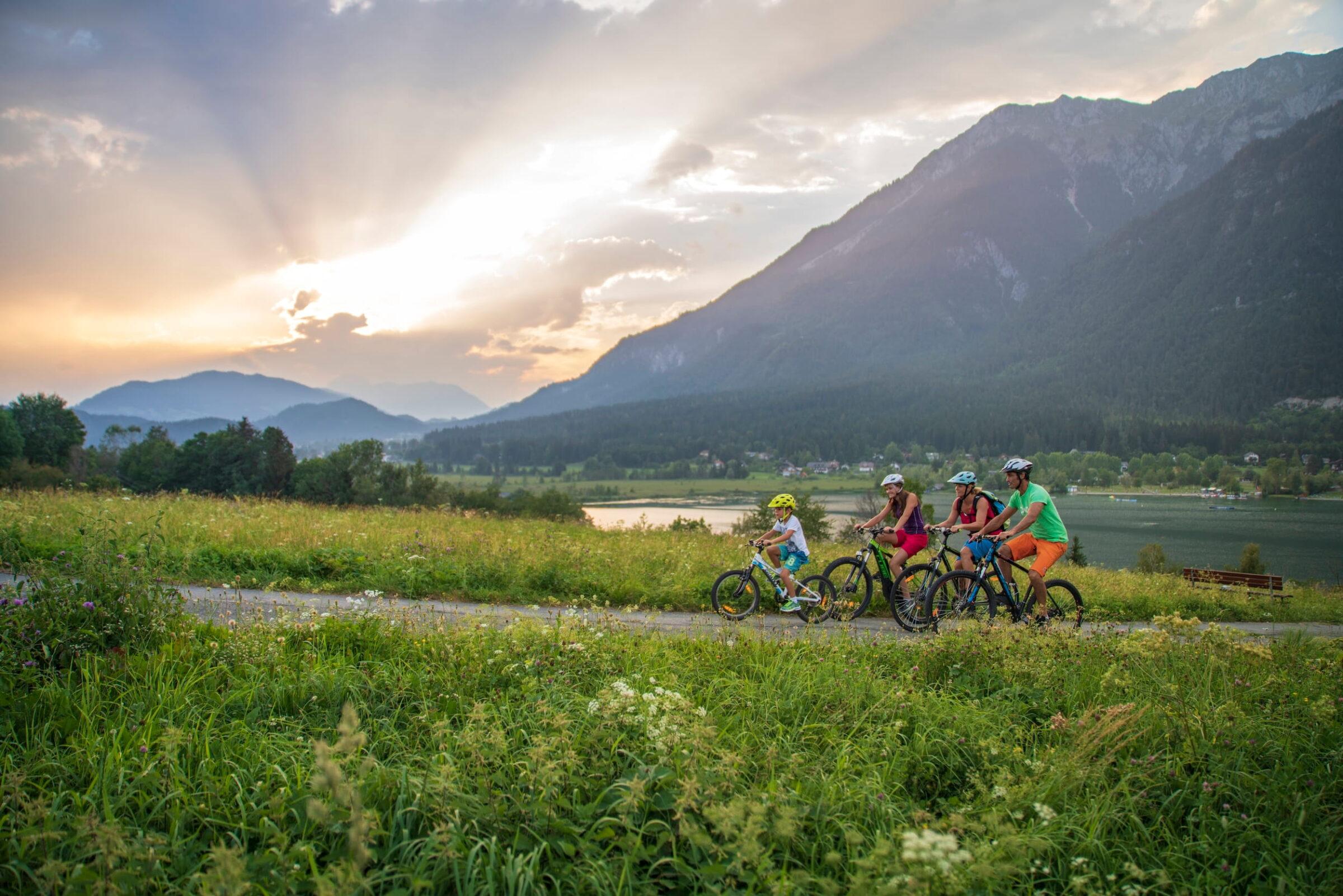 A family of four cycles along a picturesque path through lush greenery beside a lake in Carinthia, Austria, at sunset, with majestic mountains towering in the background. The mood is cheerful and happy.
